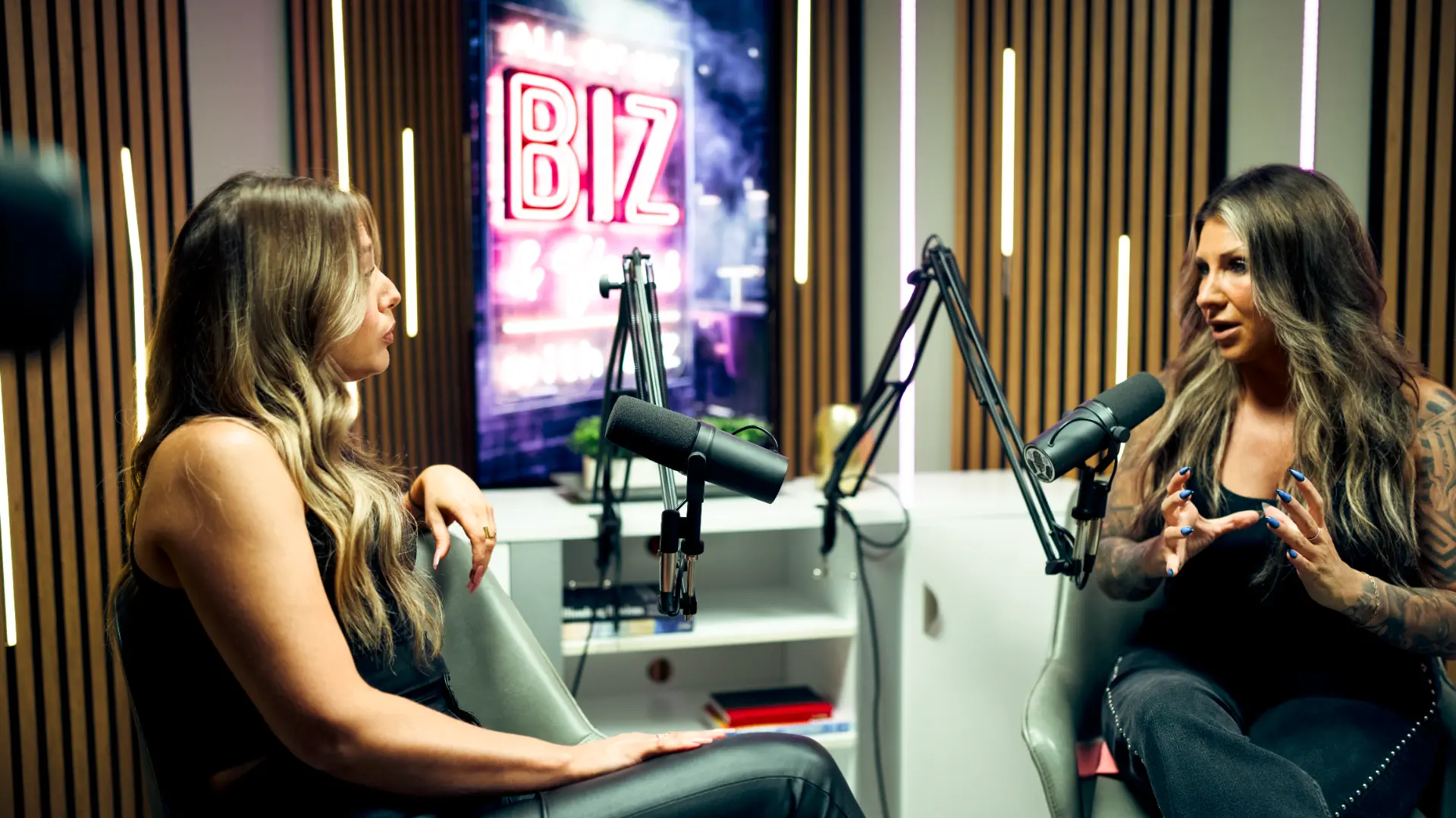 Two hosts in conversation at a podcast studio with a neon Biz sign and boom microphones