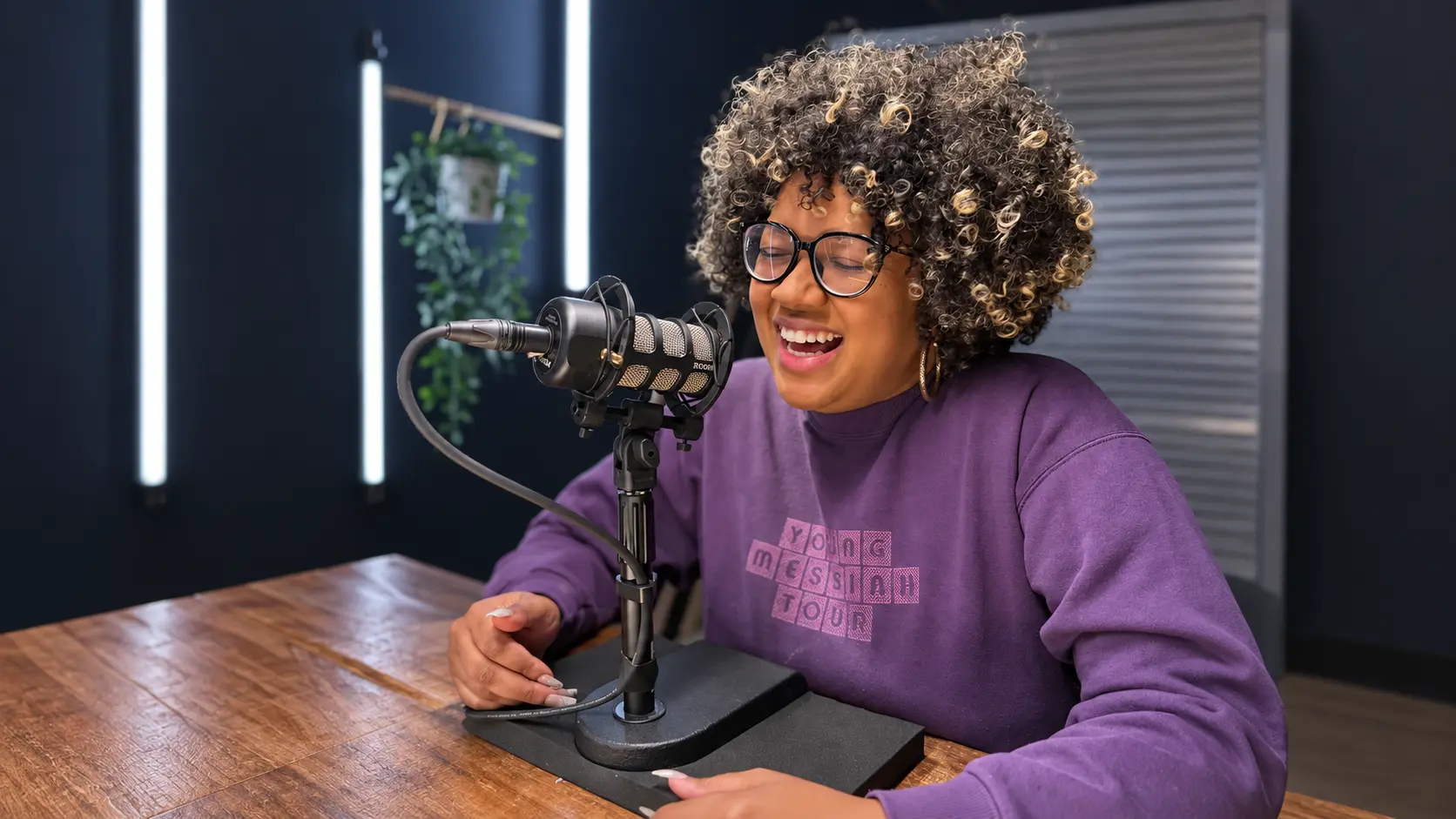 Guest smiling behind a boom microphone in a podcast studio, wearing a purple sweater